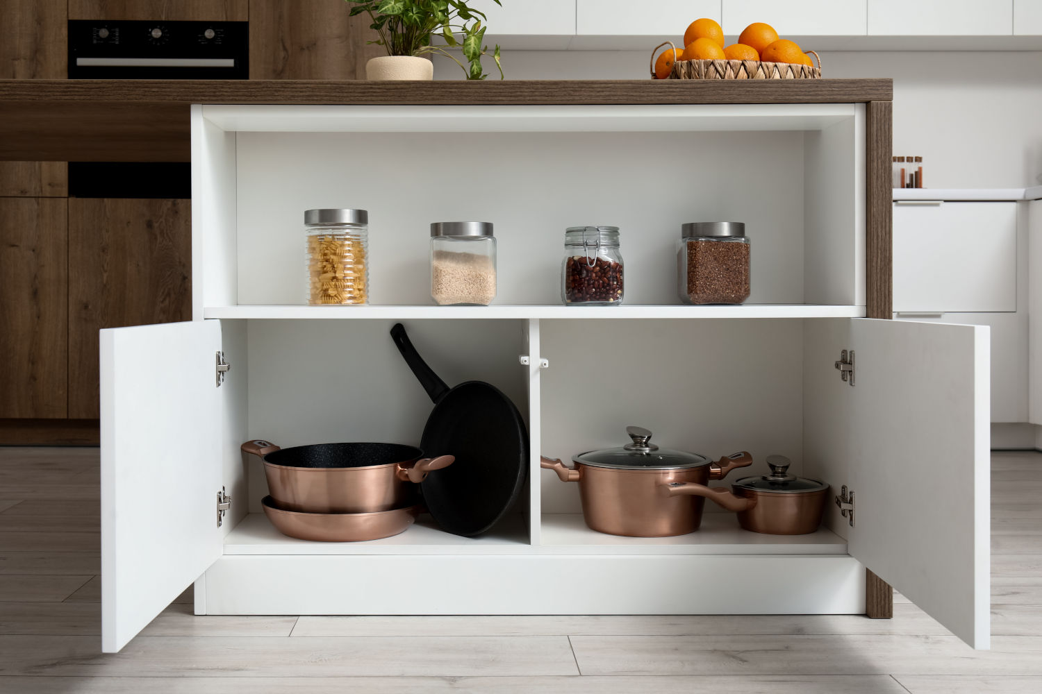 Open white shallow base cabinet with copper cookware and dry pantry goods in jars, set in a modern kitchen with light floors and minimalist decor.